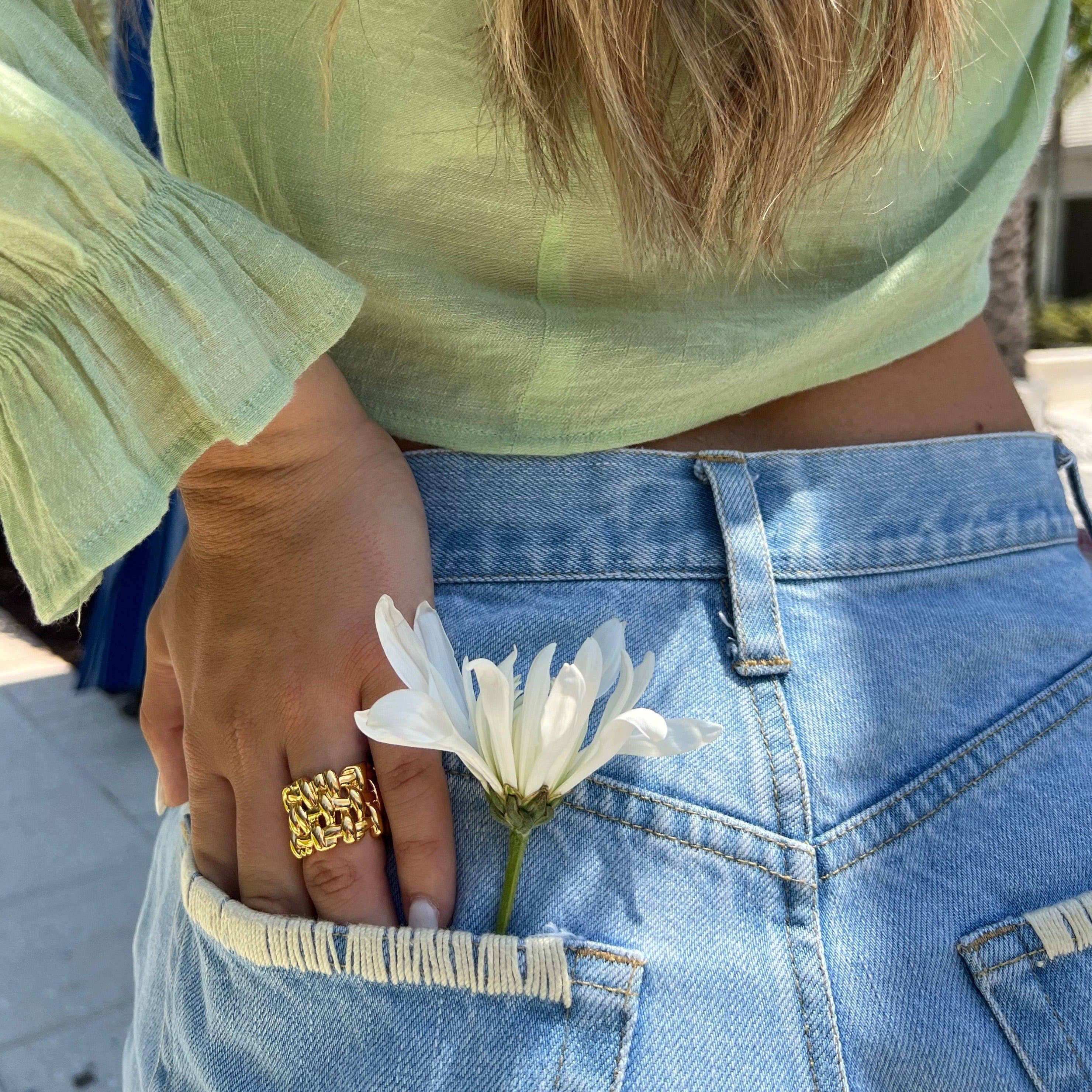 Person wearing a green top and blue jeans with a white flower in their pocket, using a gold woven ring.