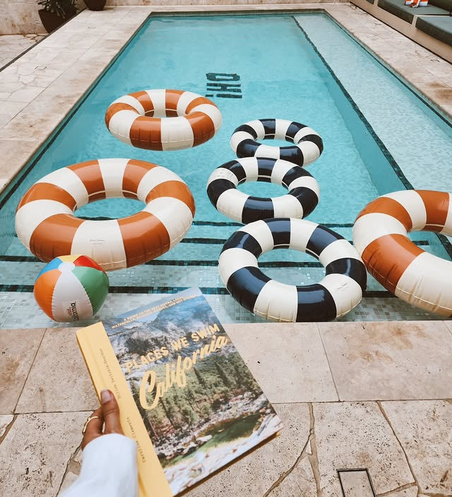 Colorful pool floats and a beach ball on a pool deck with a book titled 'California' held by a person.