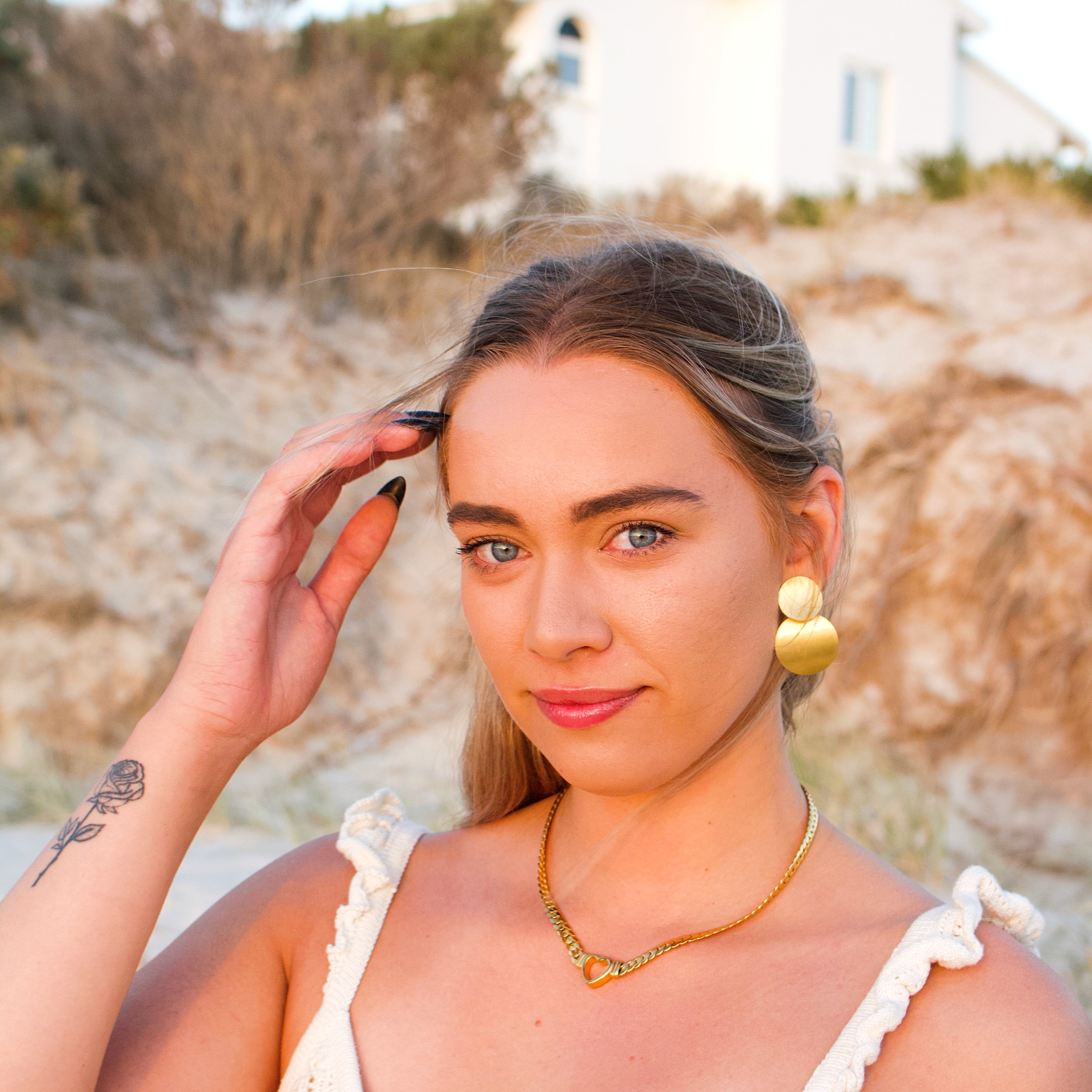 Woman with a tattoo on her arm, wearing a white top and gold jewelry, standing outdoors. 