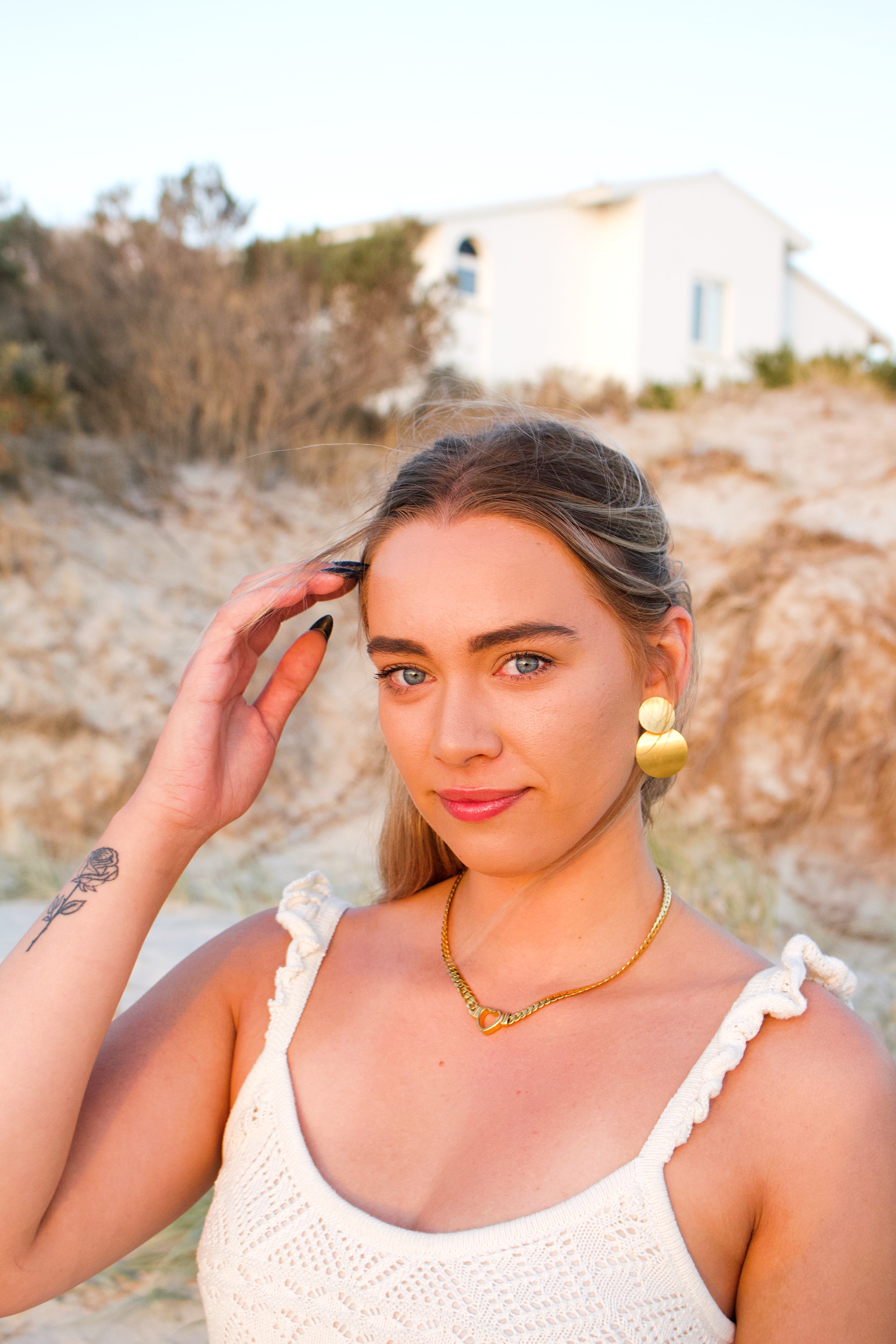 Woman with a tattoo on her arm, wearing a white top and gold jewelry, standing outdoors. 